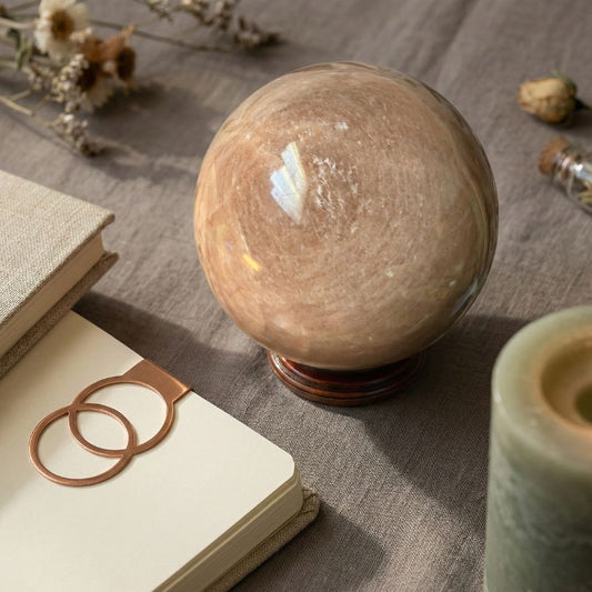Flashy Peach Moonstone Sphere — Nurturing & Balancing crystal placed on linen with books and candles, perfect for spiritual decor or meditation.