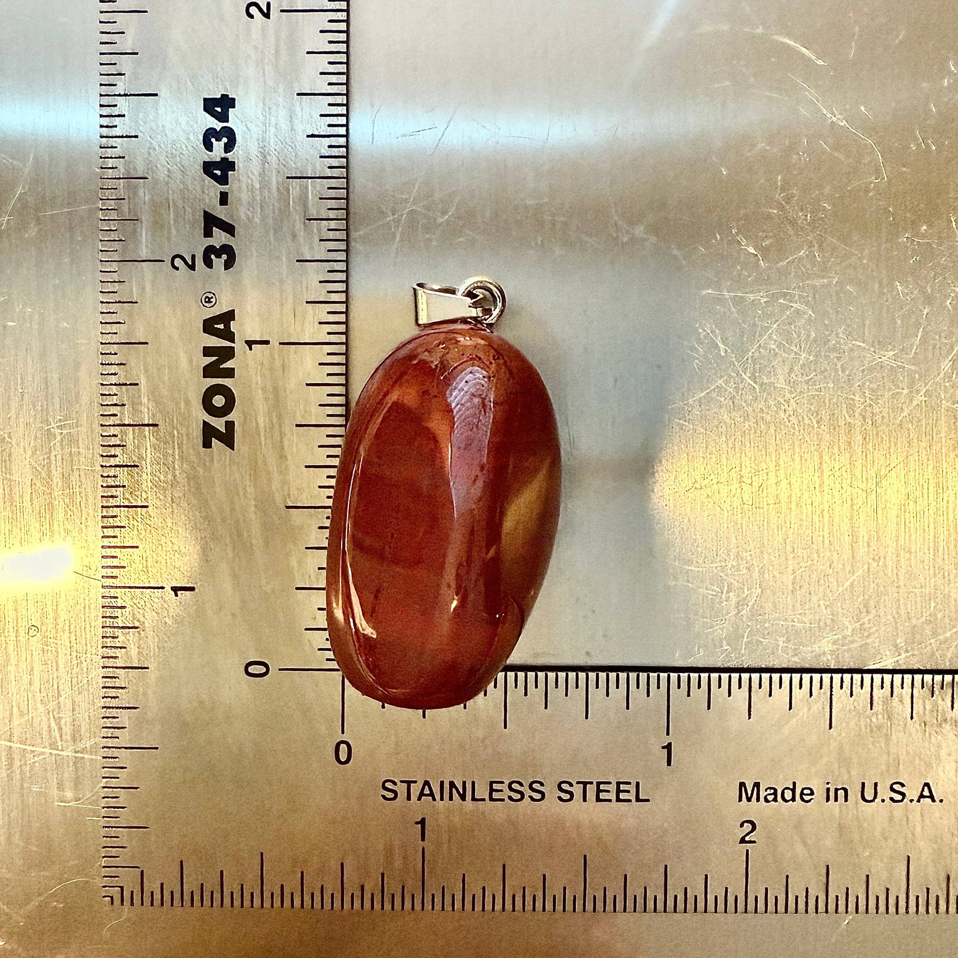 Oval-shaped natural red jasper tumble pendant with silver bale, polished surface, displayed next to a stainless steel ruler for scale.