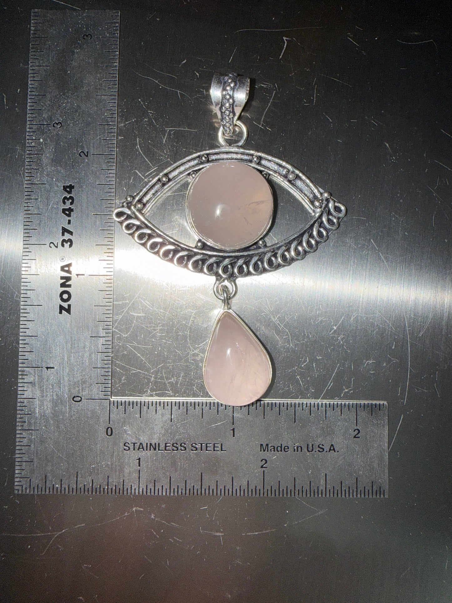 Rose Quartz Evil Eye pendant with a rose quartz drop in a silver setting on a metallic surface with ruler for scale.