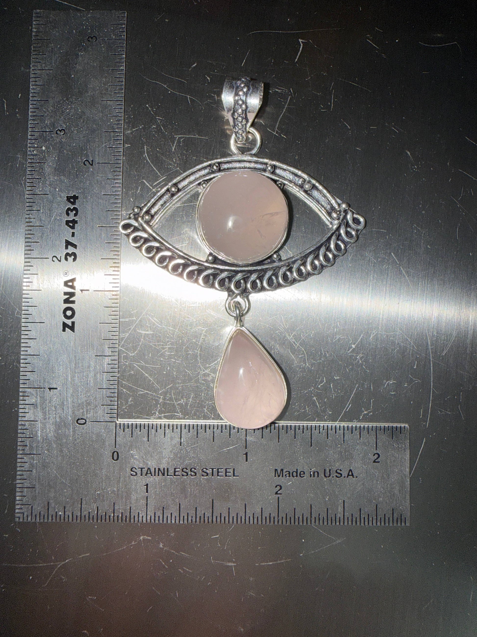 Rose Quartz Evil Eye pendant with a rose quartz drop in a silver setting on a metallic surface with ruler for scale.