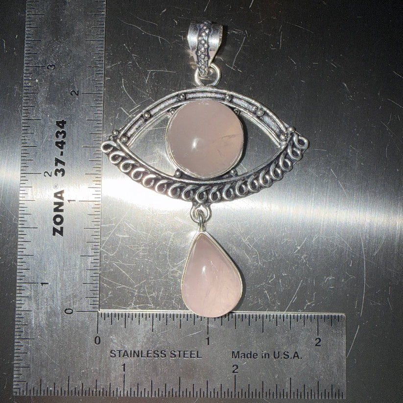 Rose Quartz Evil Eye pendant with a rose quartz drop in a silver setting on a metallic surface with ruler for scale.