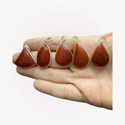 Hand holding five Carnelian Classic Pendants, each with unique red-orange tones and silver bezel, perfect for crystal jewelry lovers.