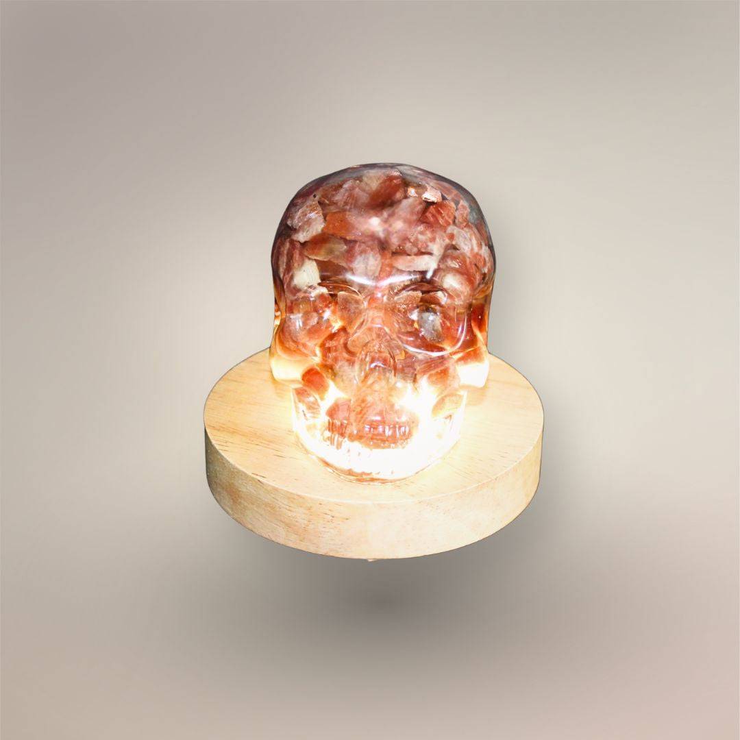 Illuminated front view of the Confetti Sunstone skull orgonite resting on a round wooden light base.
