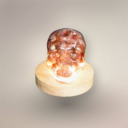 Illuminated front view of the Confetti Sunstone skull orgonite resting on a round wooden light base.