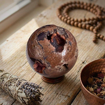 Red Fox Agate Sphere (346g) displayed on wooden table with sage and beads, showcasing its grounding energy and natural hematite-lined cavities.