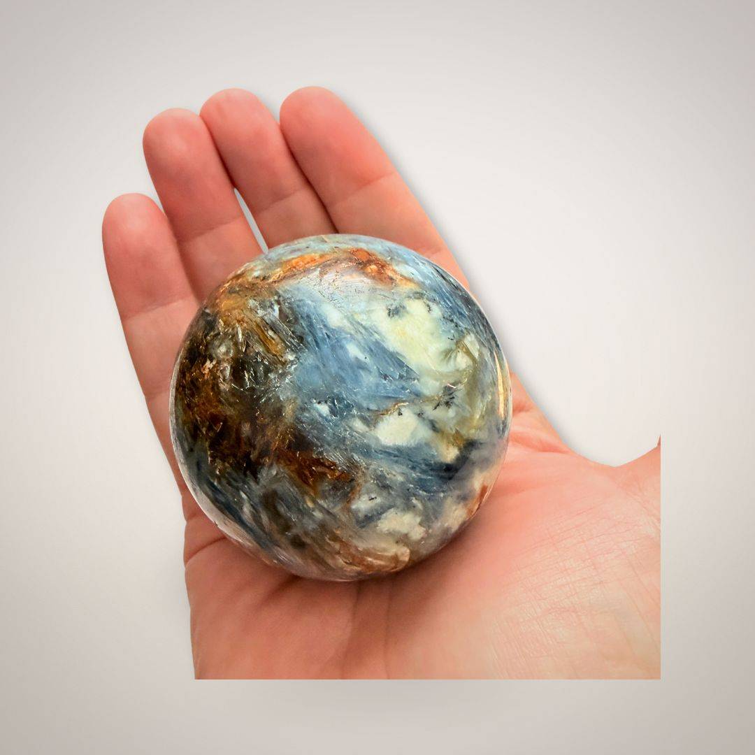 Hand holding Blue Kyanite & Quartz Sphere 61mm showcasing its polished surface and vibrant crystal inclusions for healing and energy alignment.