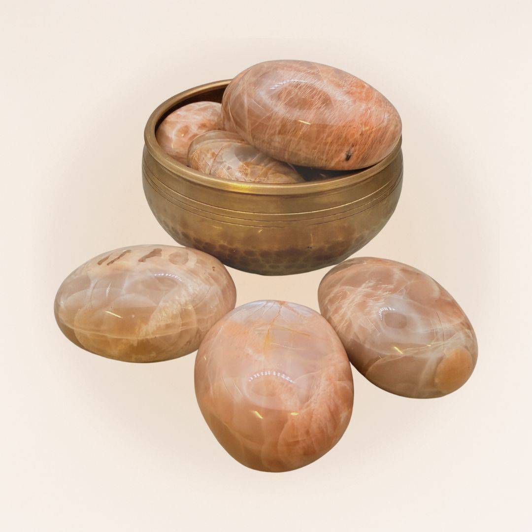 Collection of Medium Peach Moonstone Palm Stones in a brass bowl, polished crystals for calm, intuition, and emotional healing.