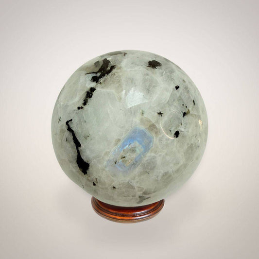 Rainbow Moonstone Sphere 50mm with black tourmaline inclusions on wooden stand, ideal for intuition and emotional balance.
