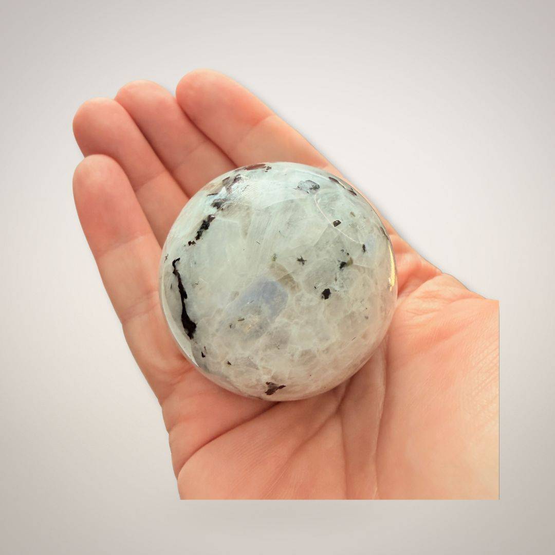 Hand holding a 50mm Rainbow Moonstone Sphere showcasing its size and natural inclusions, ideal for emotional healing and insight.
