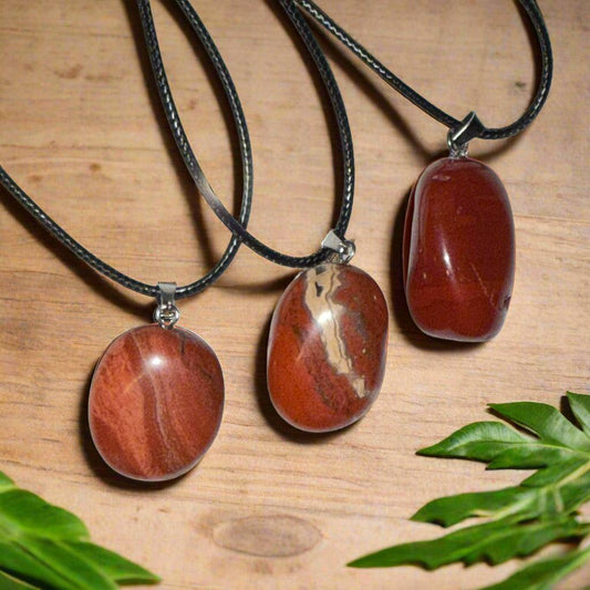 Three red jasper tumble pendants on black waxed cotton cords, showing natural variations in red, brown, and cream banding.