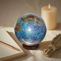 Trinity Stone Sphere 458g with Fuchsite, Kyanite, and Garnet on wooden stand beside candle, notebook, and sage bundle for spiritual decor.