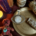 Rainbow Moonstone Evil Eye Pendant on brass tray with sage bundles and candles, symbolizing protection and intuition in spiritual jewelry.