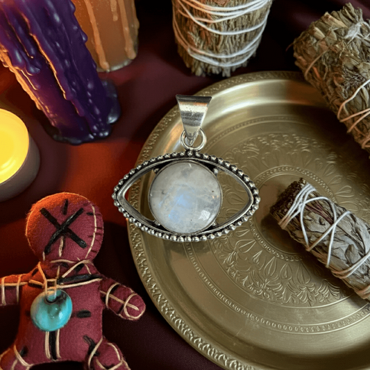 Rainbow Moonstone Evil Eye Pendant on brass tray with sage bundles and candles, symbolizing protection and intuition in spiritual jewelry.