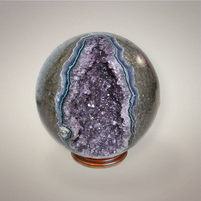 Amethyst geode sphere from Brazil with a deep purple druzy crystal opening and gray-blue agate banding, displayed on a wood stand.