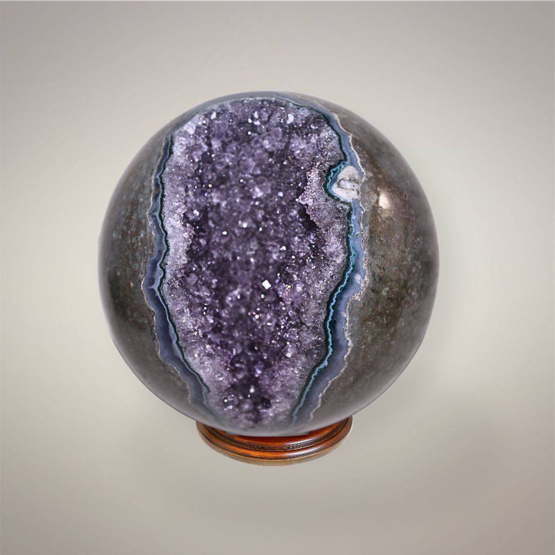 Brazilian amethyst geode sphere showing a polished gray outer surface, violet crystal-lined geode pocket, and natural blue agate banding.