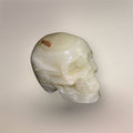 Hand-carved banded calcite skull from Mexico with cream-white layered banding, polished surface, and natural brown mineral marking.