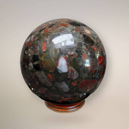 Close-up front view of a polished Conglomerate Jasper sphere on a wooden stand, showing red jasper, quartz, and dark stone fragments cemented in a natural matrix.