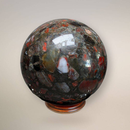 Close-up front view of a polished Conglomerate Jasper sphere on a wooden stand, showing red jasper, quartz, and dark stone fragments cemented in a natural matrix.