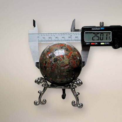 Conglomerate Jasper sphere being measured with digital calipers showing 2.501 inches, sitting on an ornate silver metal stand.