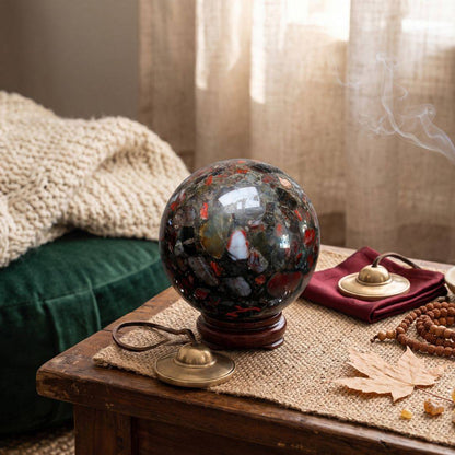 Conglomerate Jasper sphere resting on a wooden stand in a warm space alongside tingsha bells, mala beads, an autumn leaf, and drifting smoke.