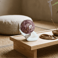 White ceramic hand stand holding a purple sphere with copper spiral on a wooden table beside incense and wooden beads.