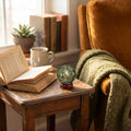 Green Aventurine orgonite sphere with copper spiral displayed on a wooden side table beside an open book, mug, plant, and cozy chair