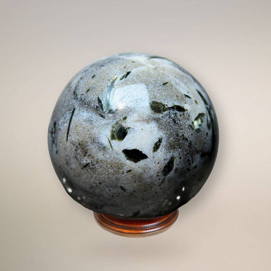 Close-up front view of a polished Green Tourmaline in Milky Quartz sphere on a wooden stand, showing dark green needle inclusions against milky white and grey quartz.