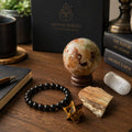 Grounded Energy Gift Set displayed on a wood desk beside a journal, fountain pen, and books, featuring a shungite bracelet, jasper sphere with wood stand, UV reactive petrified wood, tiger’s eye merkaba, and satin spar tumble.