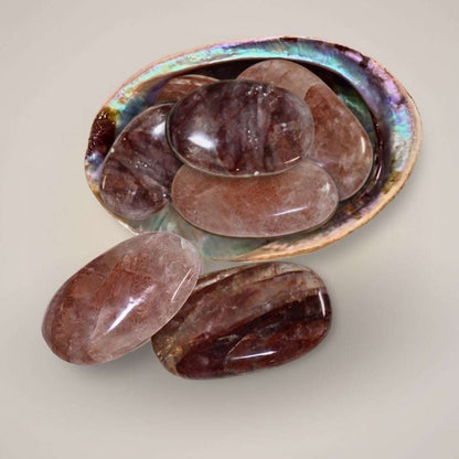 Large polished hematoid quartz palm stones from Brazil arranged in an abalone shell, showing red, rose, and smoky inclusions.