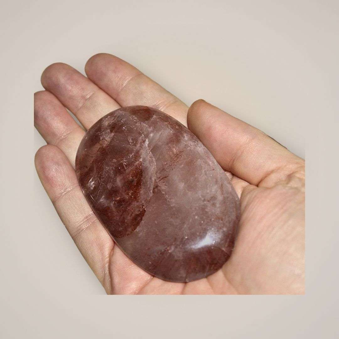 Large hematoid quartz palm stone held in hand, showing translucent quartz with warm red iron-rich inclusions.