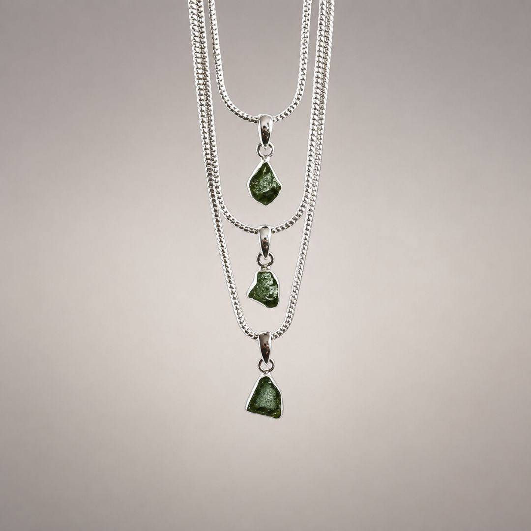 Three natural Moldavite pendants in varied irregular shapes and green tones, displayed on silver chains to show individual variation.