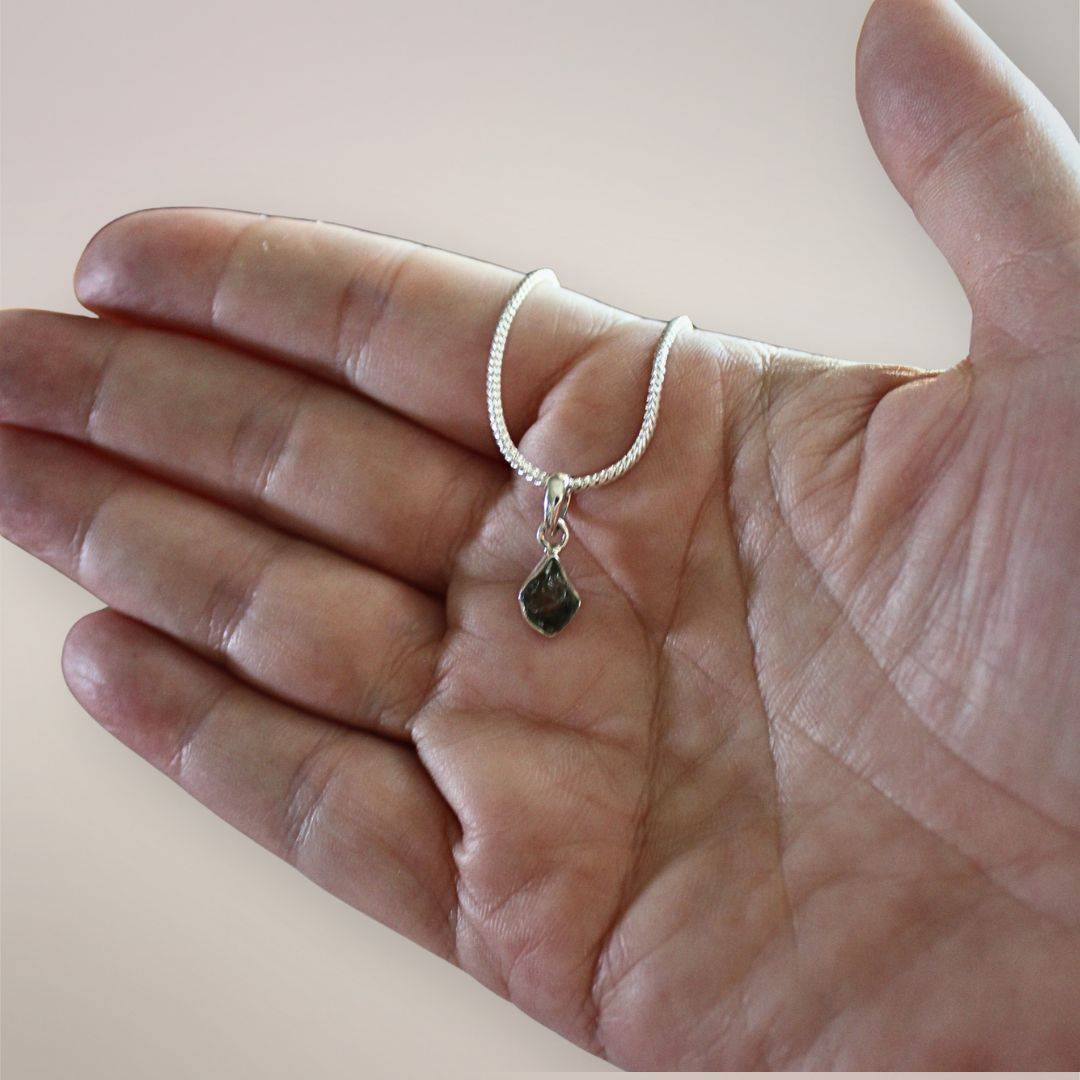 Natural Moldavite pendant resting in an open hand to show its small scale, irregular outline, and sterling silver setting.