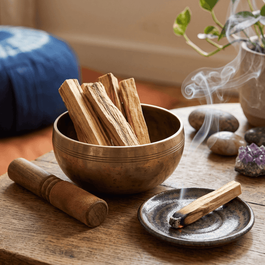 Several Palo Santo sticks resting in a brass singing bowl next to a lit, smoking stick on a small dark ceramic dish in a cozy room.