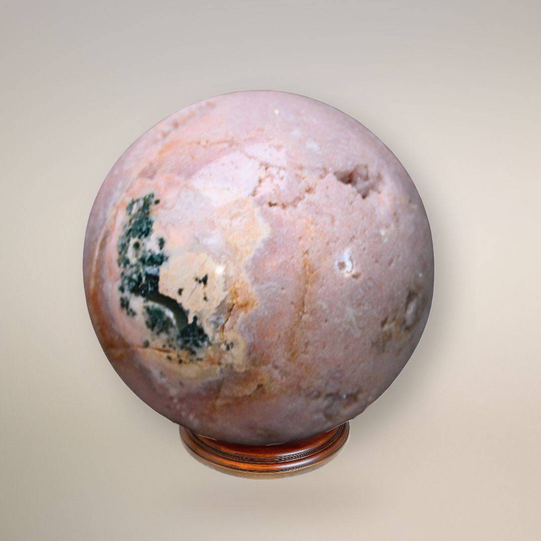Pink amethyst sphere showing pale pink coloration, an exposed crystalline geode cavity, and dark green inclusions on a wood display base.