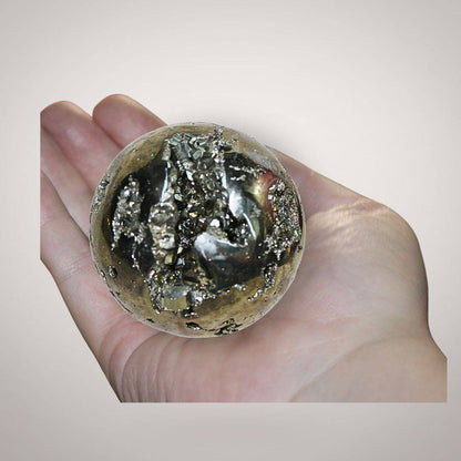 Pyrite geode sphere from Peru held in hand, showing metallic golden crystal pockets, natural matrix, and approximate palm-size scale.
