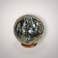 Pyrite geode sphere from Peru held in hand, showing metallic golden crystal pockets, natural matrix, and approximate palm-size scale.