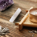 Selenite stick resting on a slatted wood surface next to dried botanical leaves and a matte vessel.