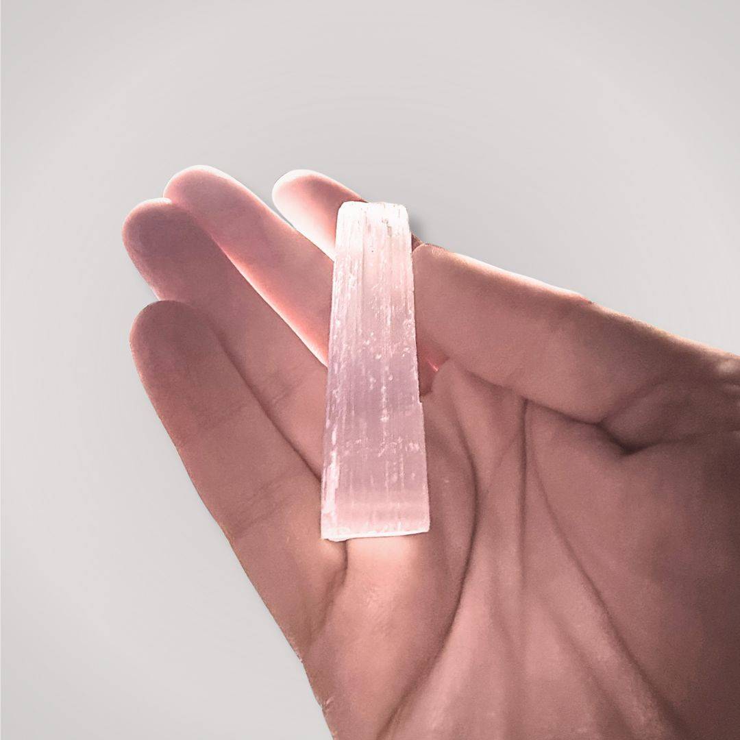 Hand holding a small unpolished selenite stick in natural light to demonstrate its 2.5-inch scale and translucent striations.