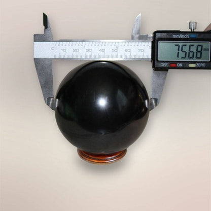 Shungite sphere measured with digital calipers showing approximately 75.68mm diameter on wooden display stand