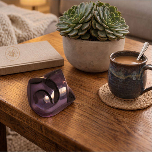 Velvet Rainbow Obsidian freeform displayed on a wooden table beside a ceramic mug and succulent, showing its polished sculptural form and soft purple coloration in direct indoor light.