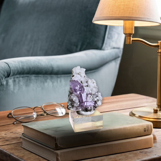 Lifestyle view showing the 2-inch amethyst specimen on a clear acrylic base resting on a stack of books for realistic scale in a calm environment.